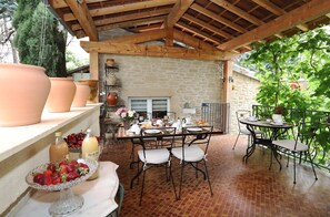 A covered patio with a stone wall, wooden beams, a table set for dining, and potted plants.