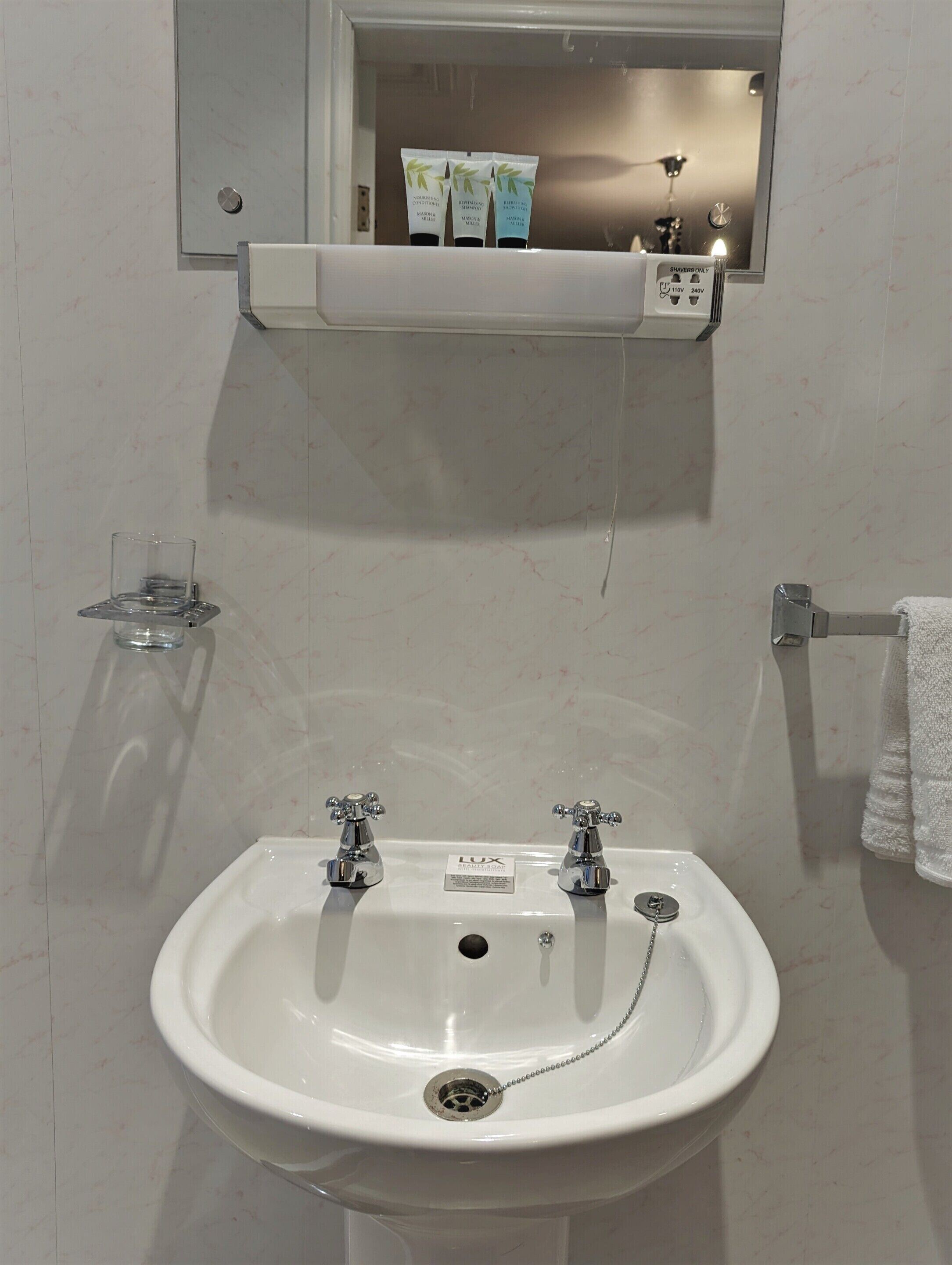 A bathroom with a white sink, two faucets, a mirror above the sink, and a shelf with toiletries.