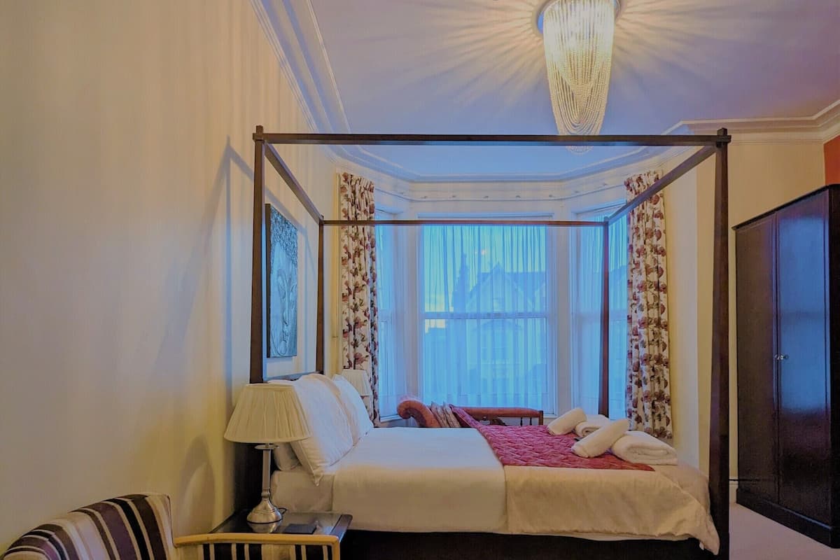 A four-poster bed with a canopy, a bedside table with a lamp, a striped armchair, and a dark wooden wardrobe.