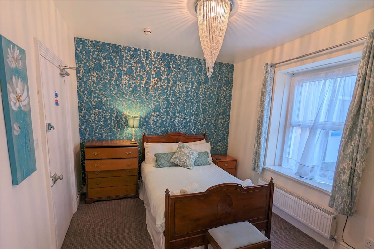 A bedroom with a wooden bed, a nightstand, a dresser, a bench, a chandelier, and a window with curtains.