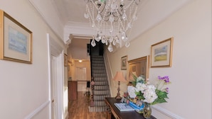 A hallway with wooden flooring, a chandelier, framed artwork, a desk with a vase of flowers, and a staircase leading to another room. - The Senarth (Llandudno)