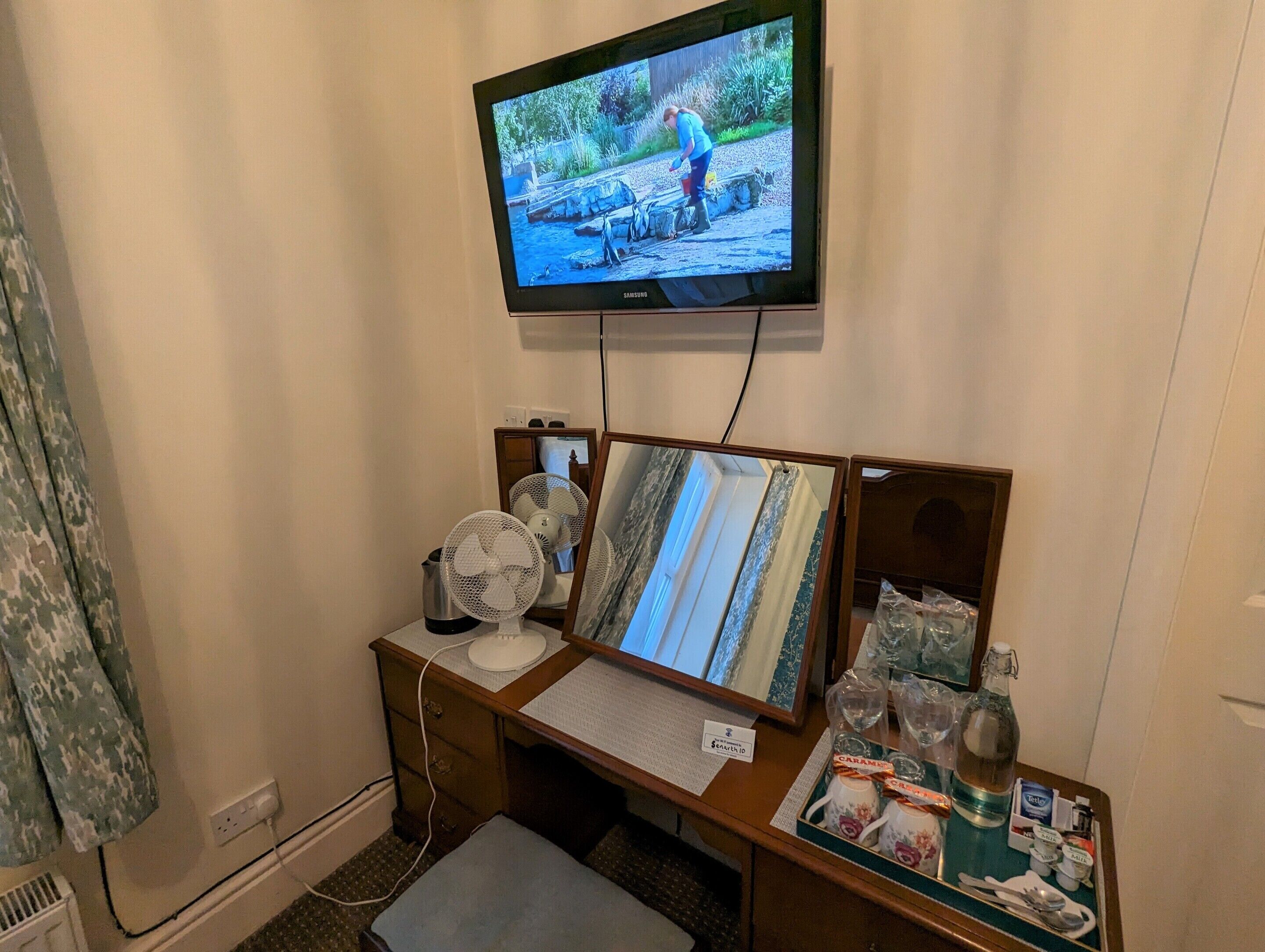 A hotel room with a television mounted on the wall, a desk with a computer, a fan, and a small table with toiletries.