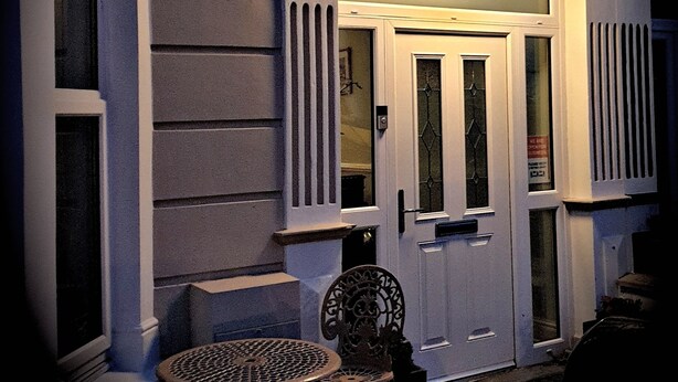 A building with a sign reading "SENARTH" above the entrance, a door with glass panels, and a small outdoor seating area with a round table and chairs. - The Senarth (Llandudno)