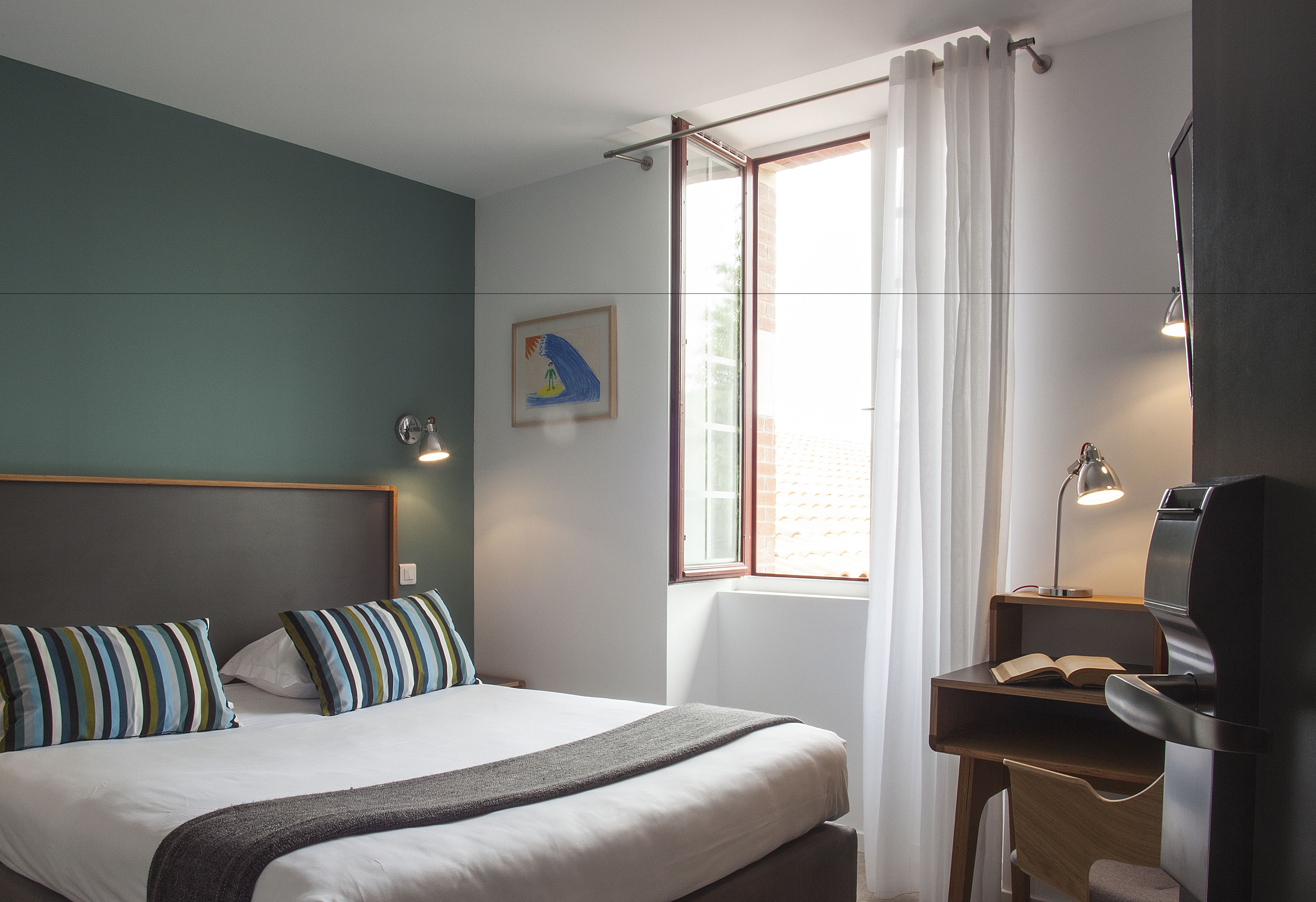 A hotel room with a bed, a desk, a chair, a window with curtains, and a painting on the wall.