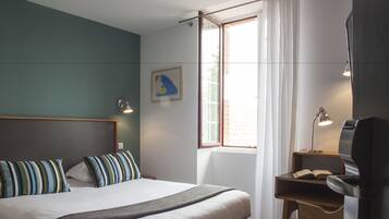 A hotel room with a bed, a desk, a chair, a window with curtains, and a painting on the wall.