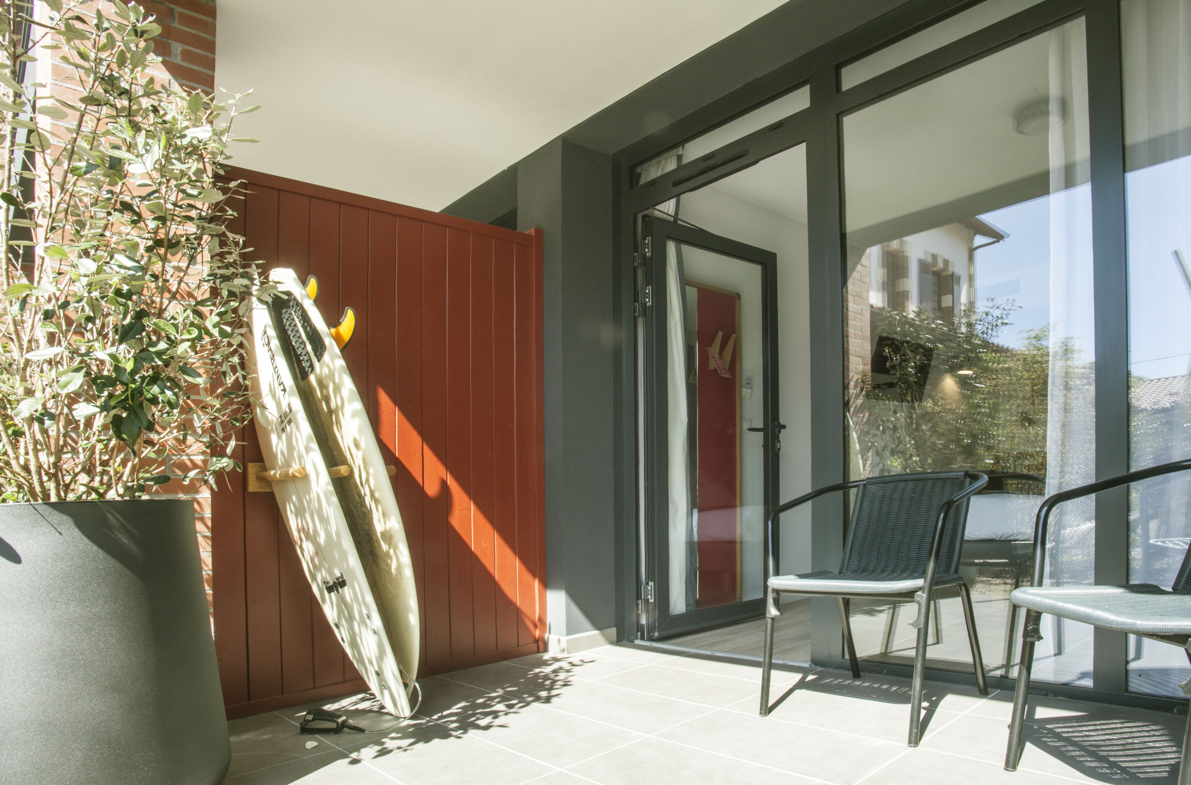 Un balcon avec une planche de surf appuyée contre une clôture en bois, deux chaises et un pot de fleurs.