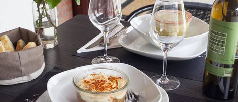 Una mesa de comedor con una botella de vino, un vaso de agua, una copa de vino blanco, un postre en un vaso y un bol de pan.