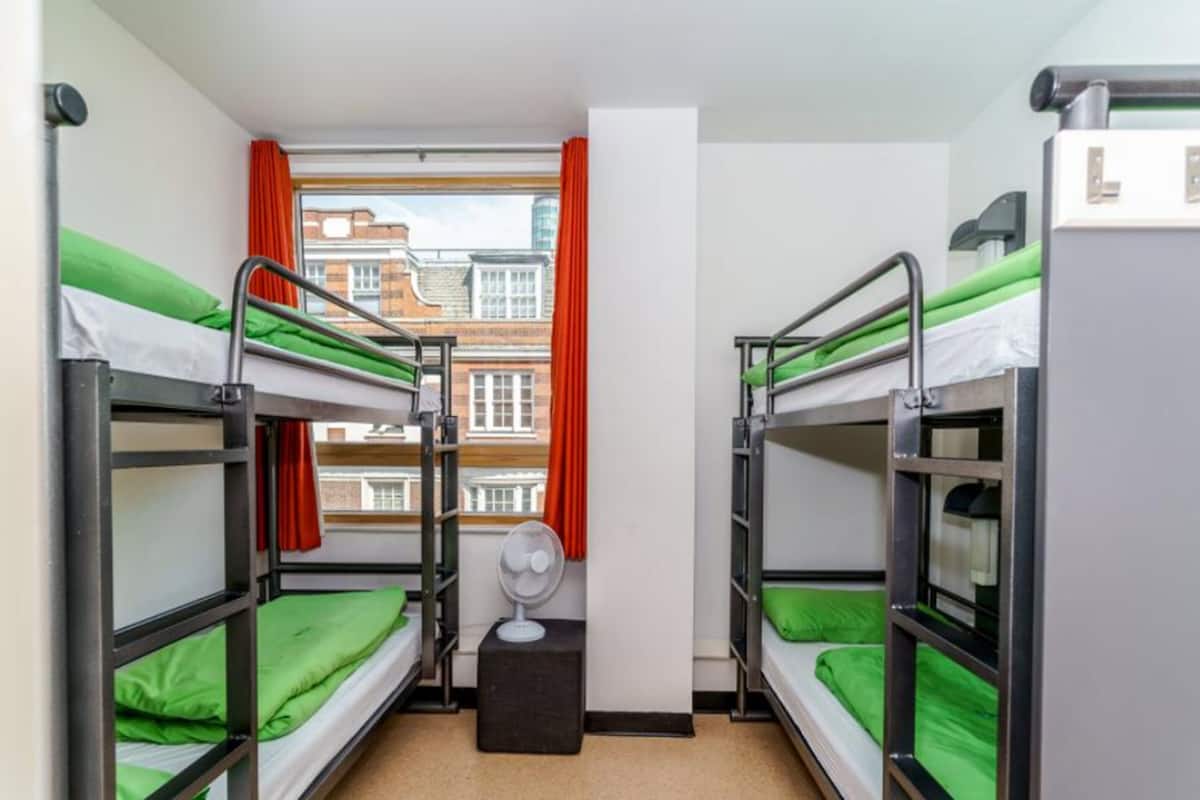 A dormitory room with bunk beds, a window with red curtains, and a fan.