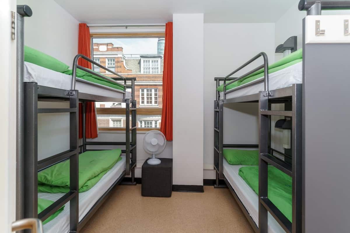 A dormitory room with bunk beds, a window with red curtains, and a standing fan.