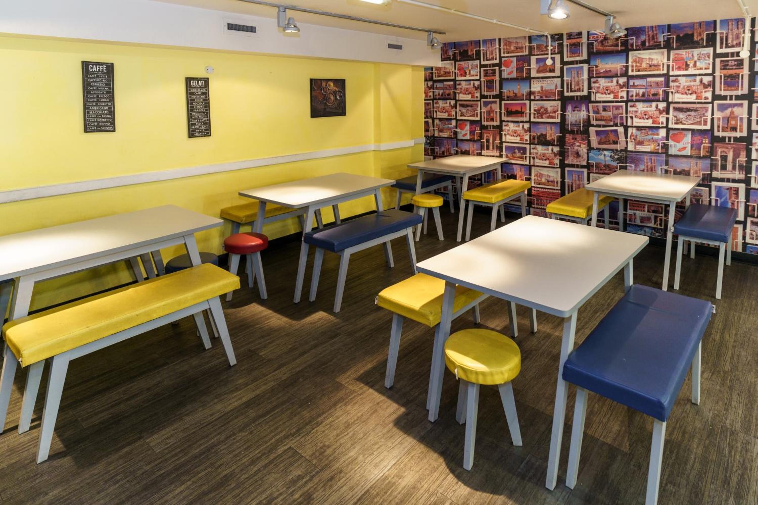 Un café avec des tables et des tabourets jaunes et bleus, un tableau affichant le menu sur le mur et un mur recouvert de cartes postales.