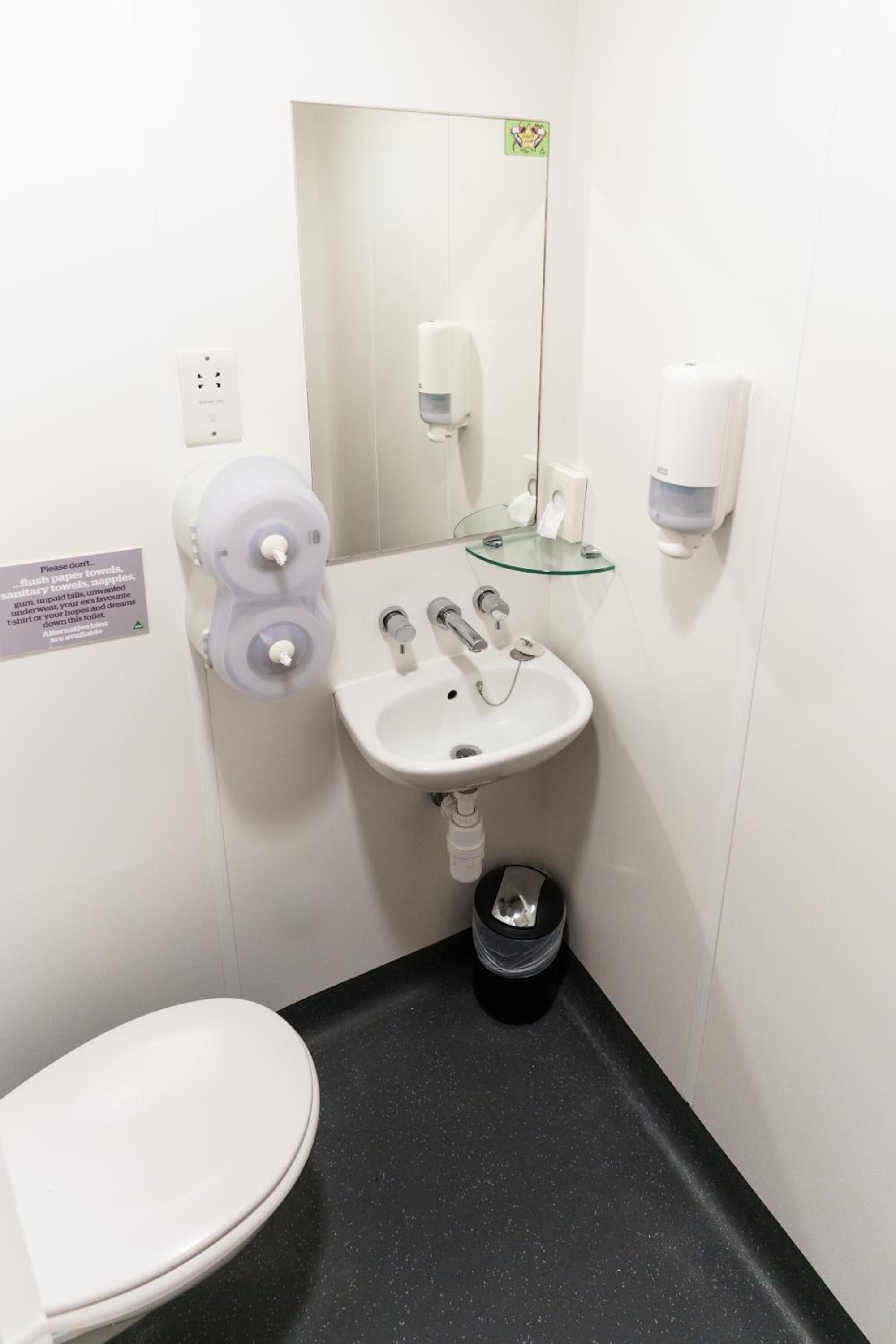 Ein kleines, modernes Badezimmer mit einem weißen Waschbecken, einem Spiegel, einer Toilette und zwei Handtrocknern.