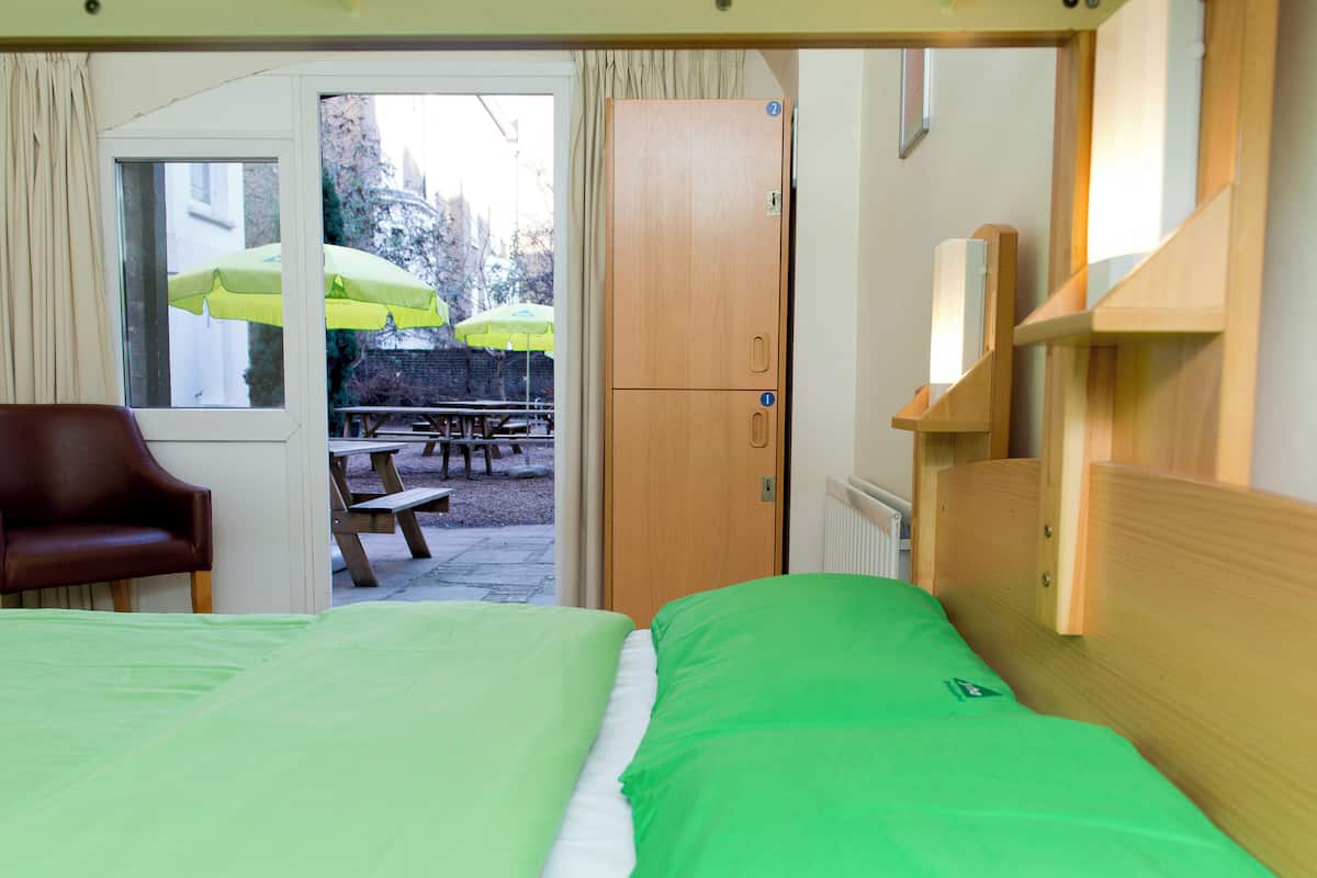 A room with a bed, a wooden wardrobe, a brown chair, and a view of an outdoor patio with picnic tables and umbrellas.