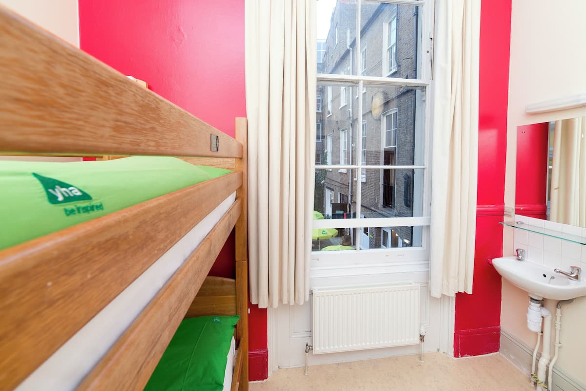 A small, brightly colored room with a wooden bunk bed, a window with curtains, a radiator, and a sink.