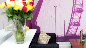 Una stanza con un murale a tema viola, un divano con cuscini decorati, un vaso con fiori e un tavolo di legno con una bottiglia.