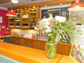 Una zona de bar con mostrador de madera, un jarrón de flores y estantes con botellas.