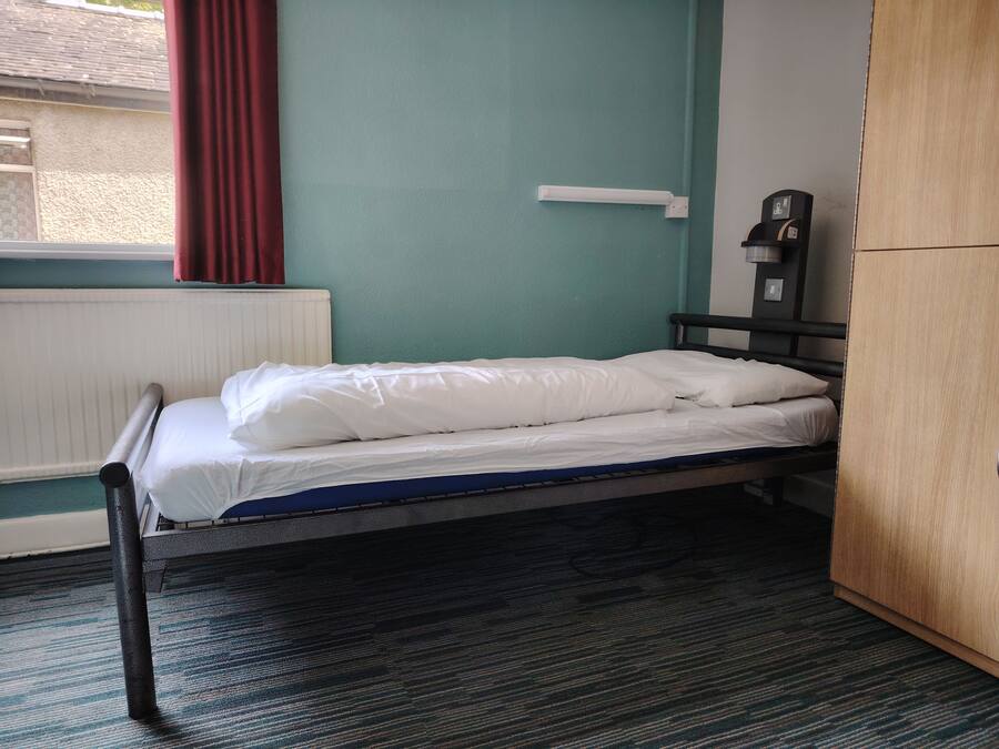 A single bed with white bedding in a room with teal walls, a radiator, and a wooden wardrobe.