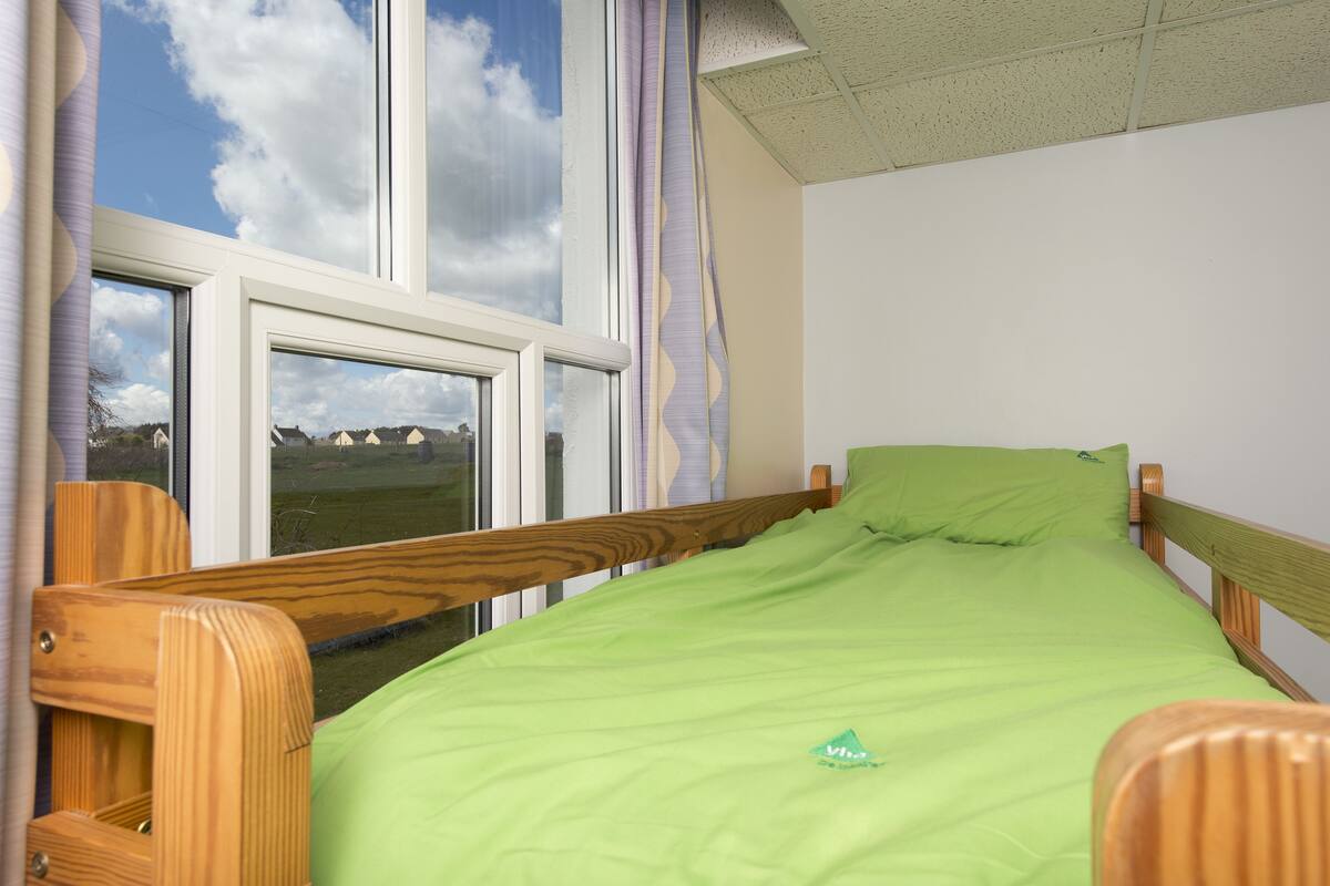 Un lit simple avec une couvre-lit verte et une tête de lit en bois, dans une pièce dotée d’une grande fenêtre et de rideaux.