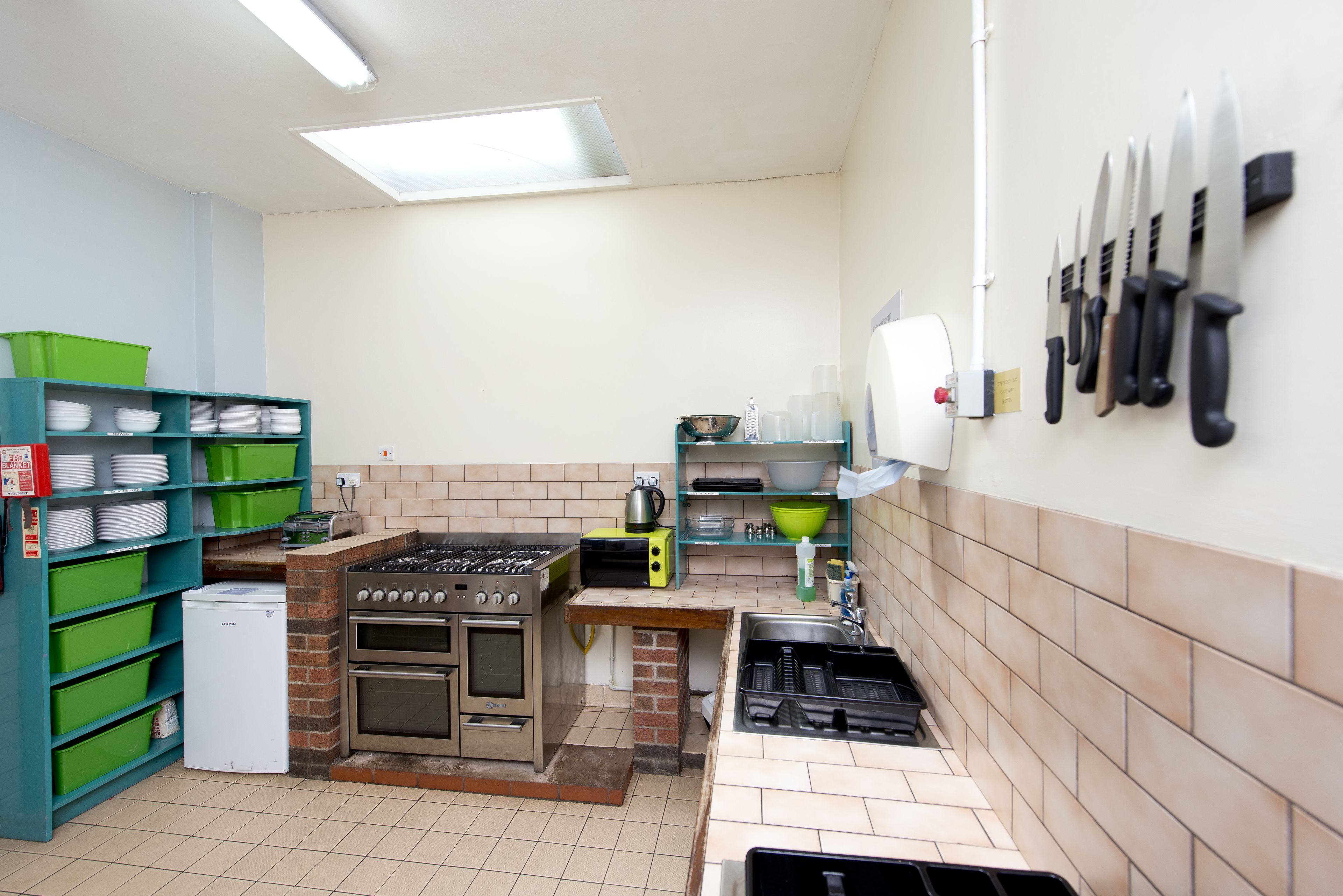 A compact kitchen with a gas stove, oven, and a small refrigerator. There are green storage bins and a sink with a faucet.