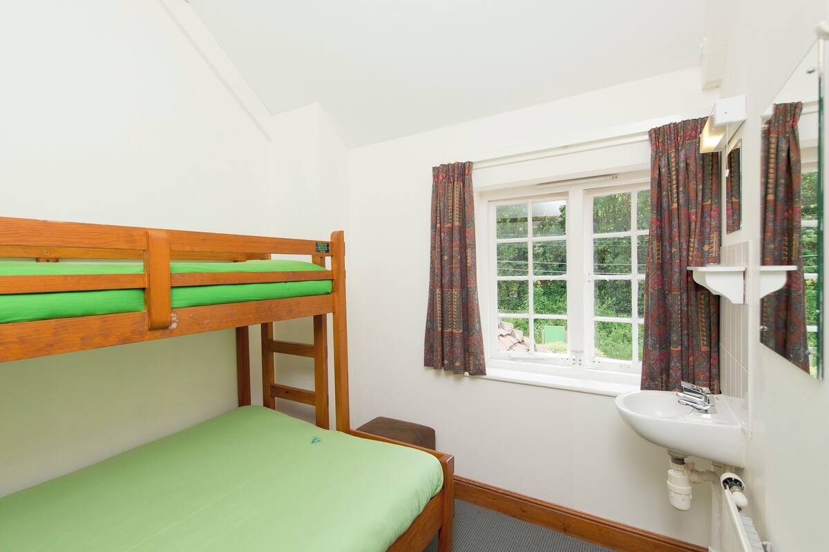 Ein Stockbett mit grüner Bettwäsche, ein kleiner Nachttisch, ein Fenster mit gemusterten Vorhängen und ein weißes Waschbecken mit Spiegel.