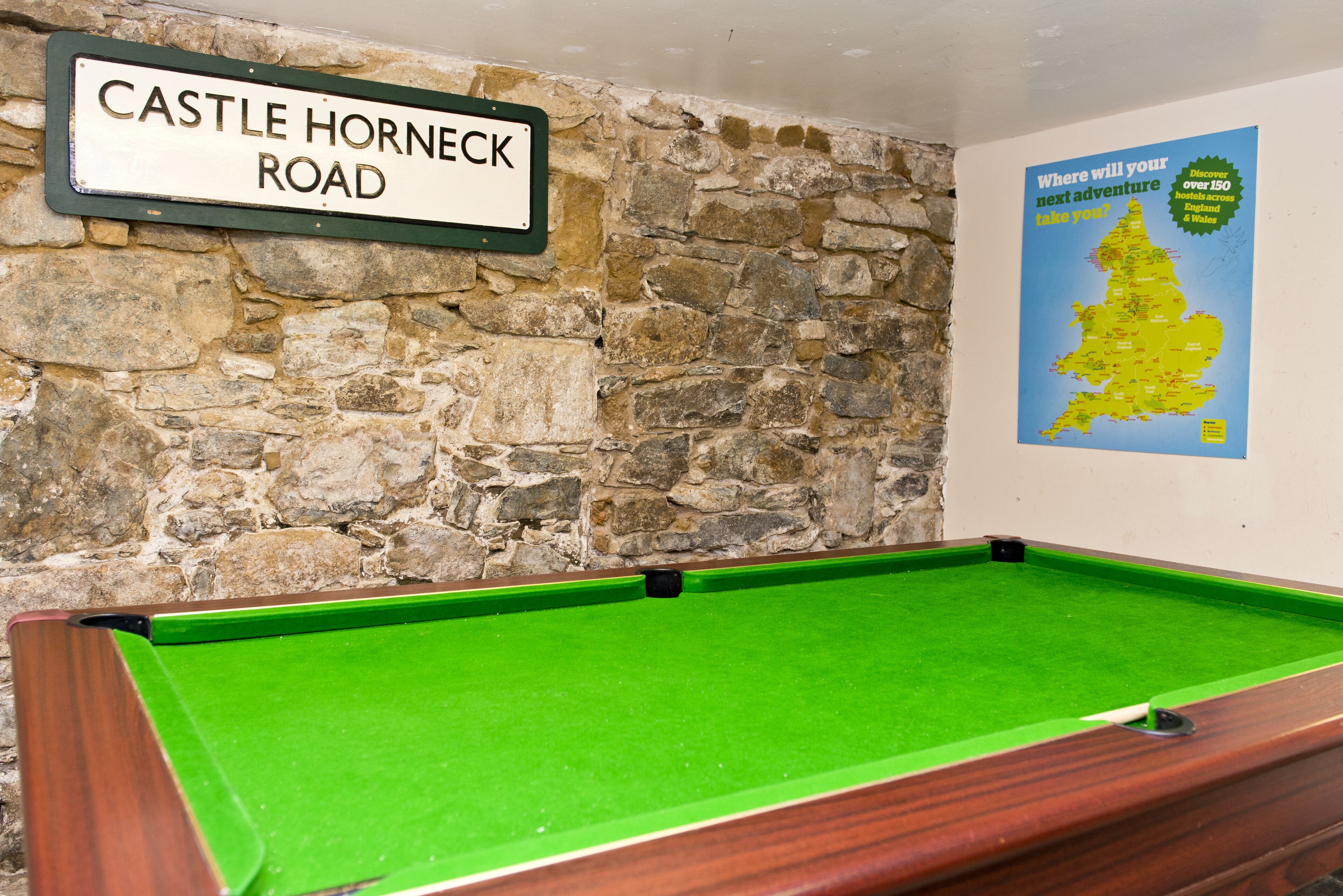 Una mesa de billar con superficie de fieltro verde, una pared de piedra y un cartel que dice "CASTLE HORNECK ROAD".