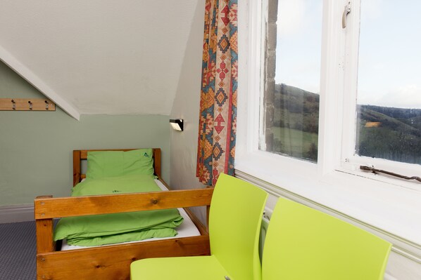 A small, single-bed room with a window, a wooden headboard, and a green chair.