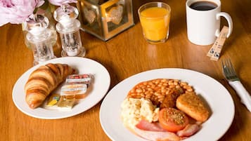 A breakfast setting with croissants, butter, jam, a plate of beans, eggs, tomatoes, ham, and toast, along with a coffee and orange juice.