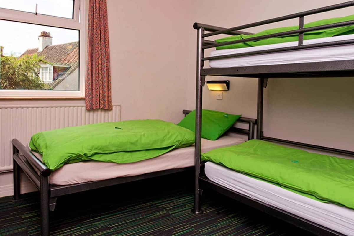 Dormitorio con literas, ropa de cama verde, una ventana con vistas a casas y un radiador.