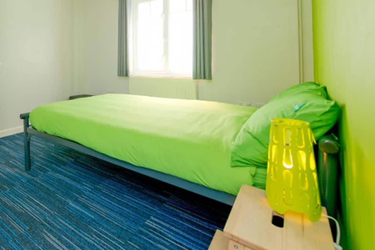 Una habitación con cama individual, colcha verde, ventana con cortinas, alfombra azul y lámpara amarilla sobre una mesita de noche de madera.