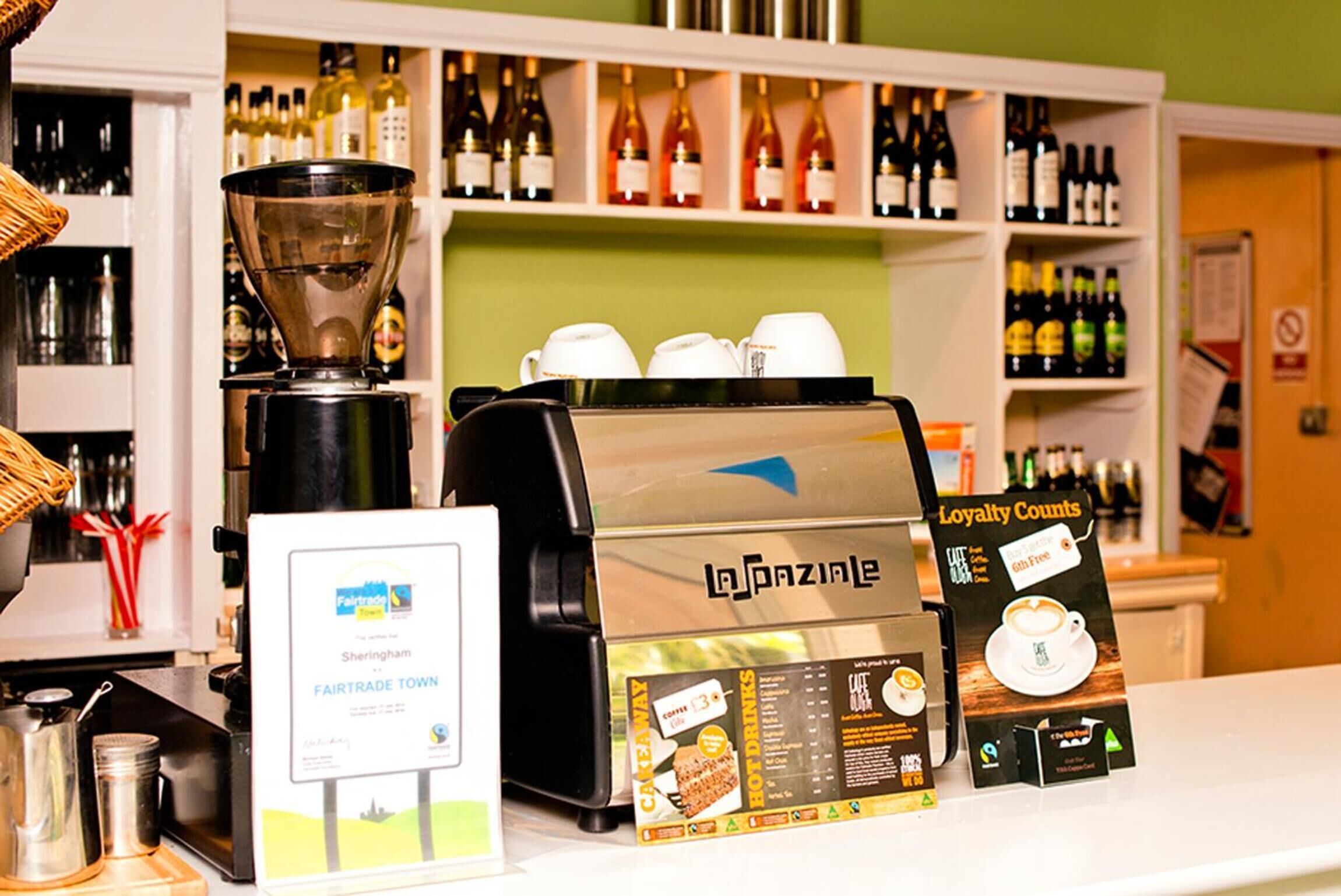 A coffee machine with a loyalty program sign, a coffee cup, and a shelf with wine bottles.