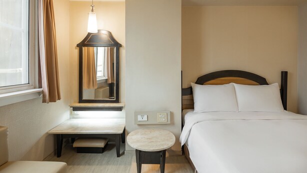 Ein Hotelzimmer mit Bett, Nachttisch, einem kleinen runden Tisch, einem Spiegel und einem Fenster mit Vorhängen.