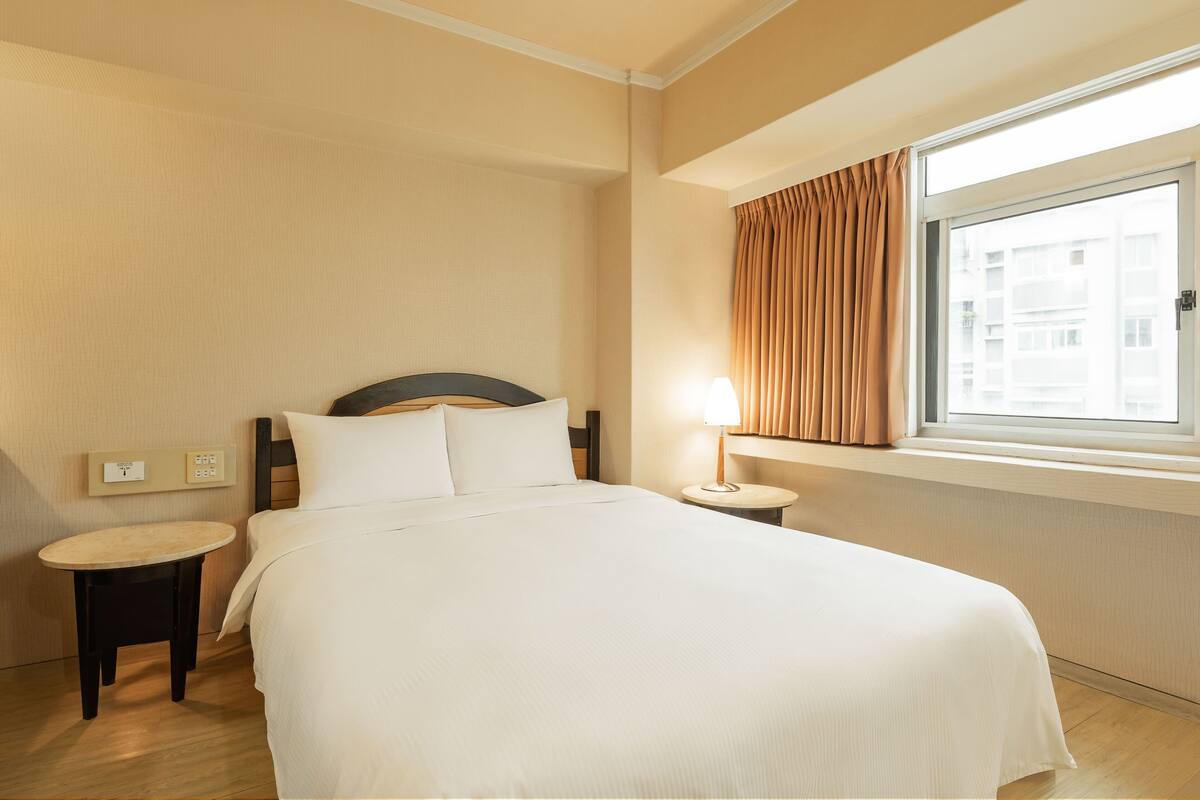 Ein Hotelzimmer mit einem großen Bett, einem Nachttisch, einer Lampe und einem Fenster mit Vorhängen.