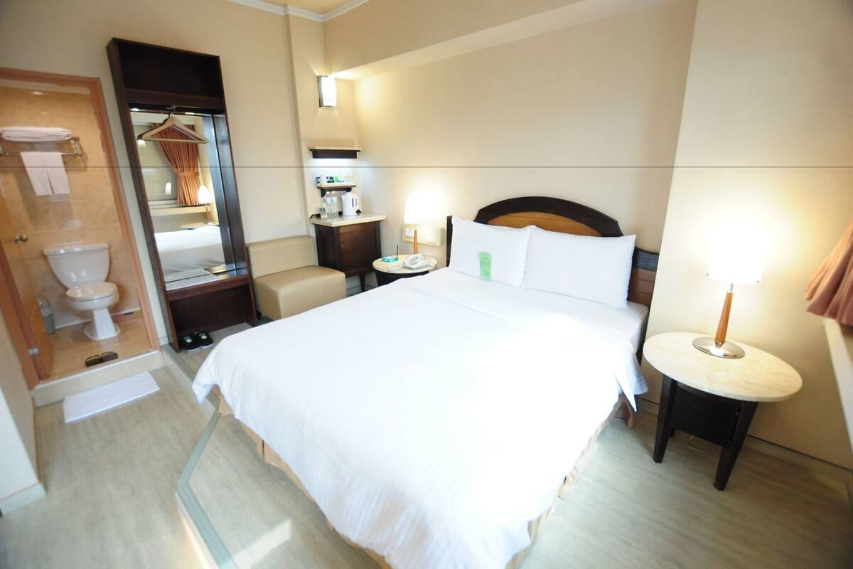 Ein Hotelzimmer mit Bett, Badezimmer mit Toilette, Spiegel und Nachttisch mit Lampe.