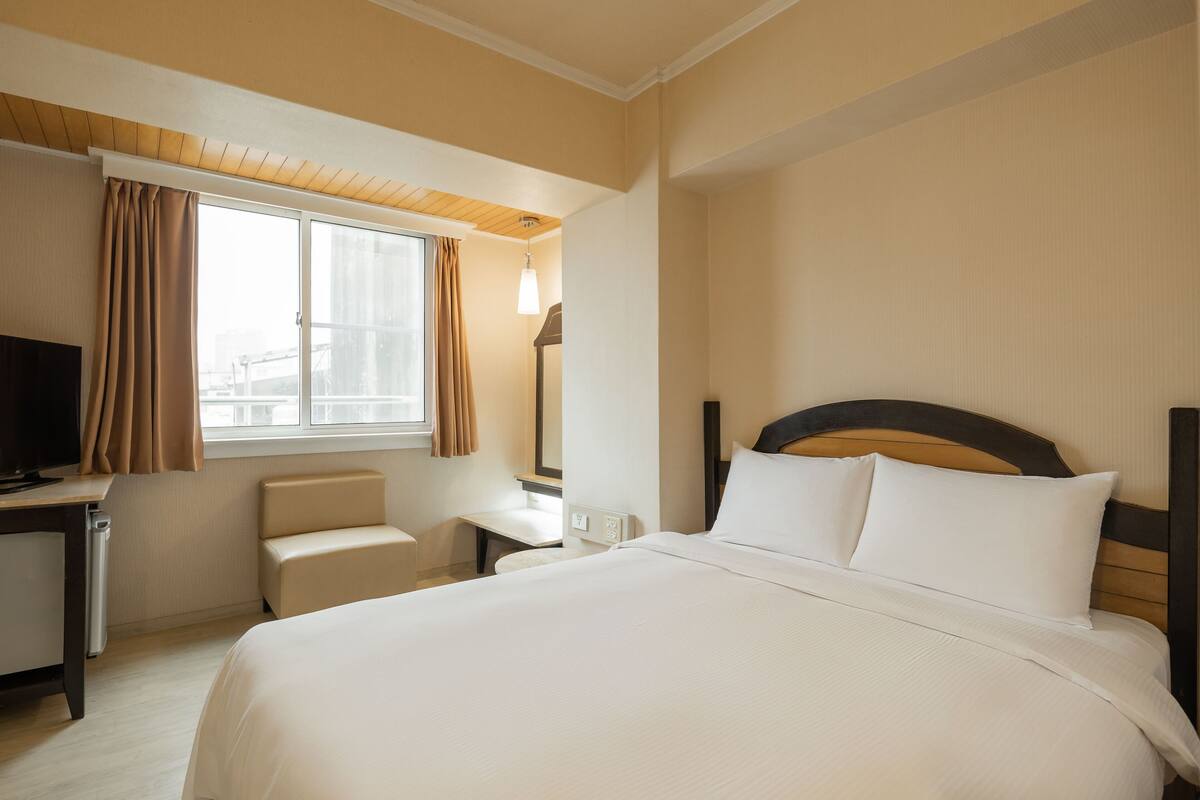 Ein Hotelzimmer mit einem großen Bett, einem Schreibtisch mit Fernseher, einem kleinen Tisch und einem Fenster mit Vorhängen.