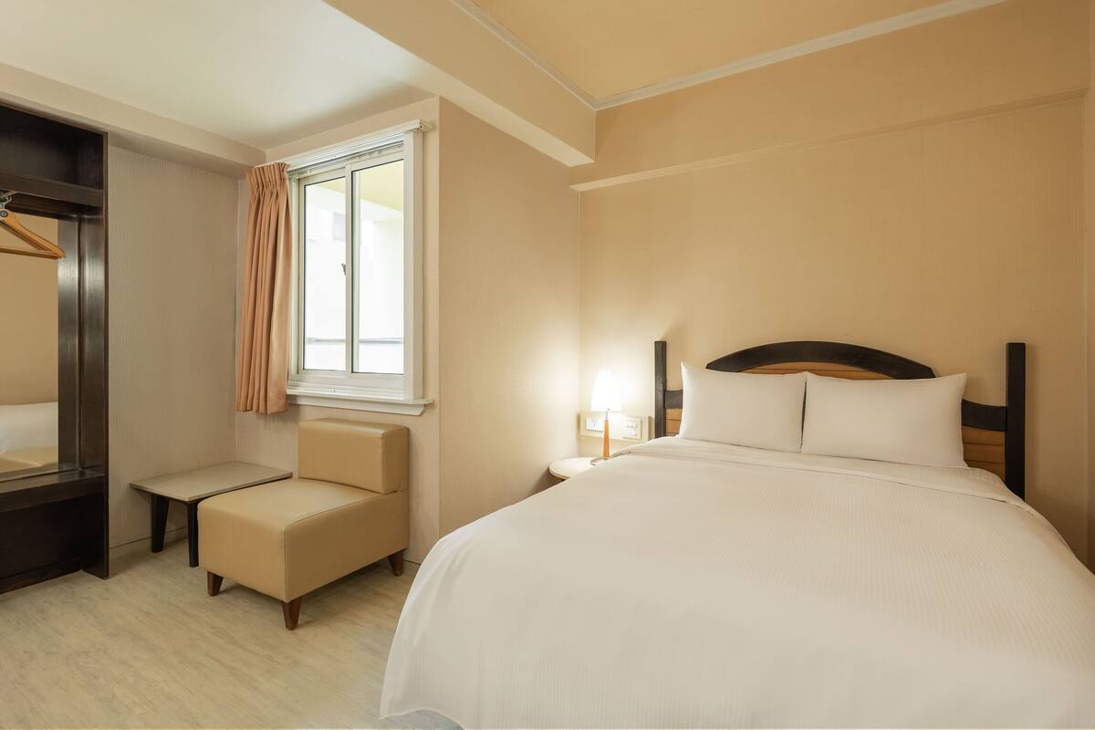 Ein Hotelzimmer mit einem großen Bett, einem Nachttisch, einem Sessel, einem Kleiderschrank und einem fenster mit Vorhängen.