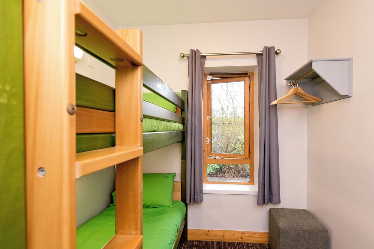 A room with bunk beds, a small window with curtains, a hanging coat rack, and a grey upholstered bench.