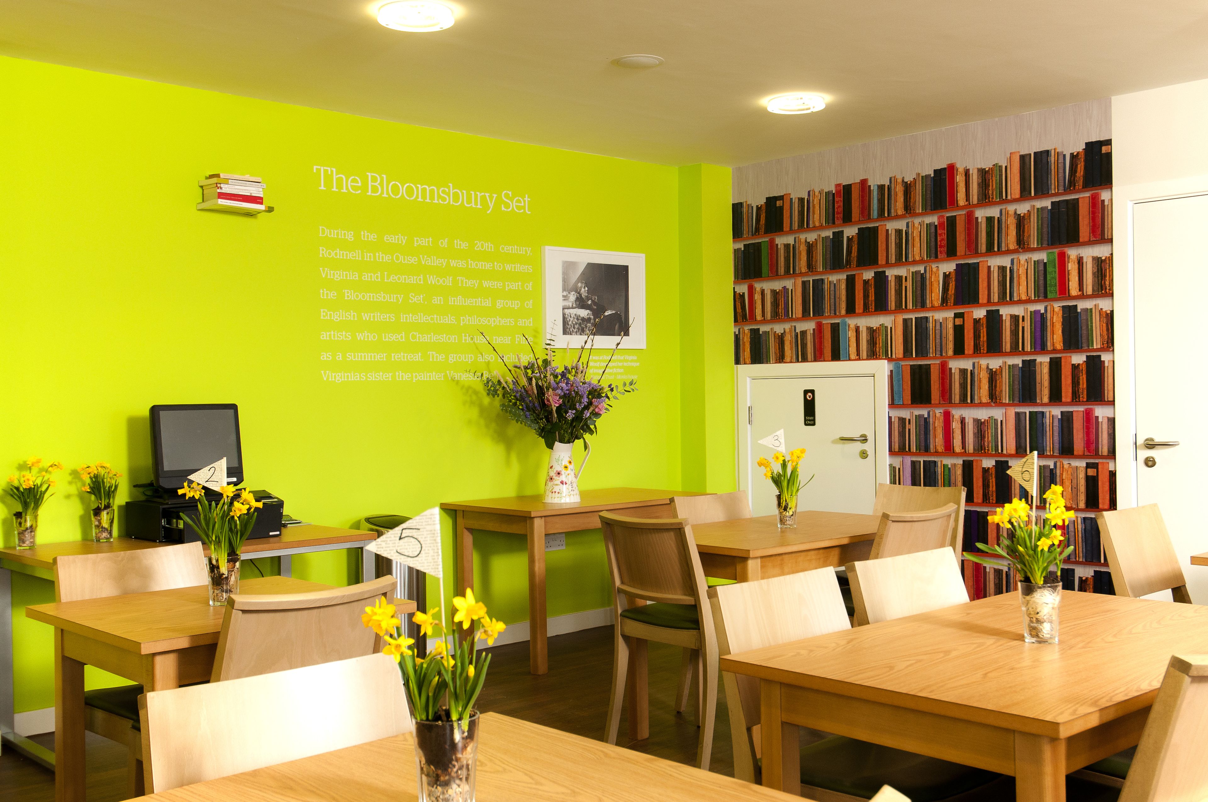 A library with green walls, multiple tables, chairs, and a bookshelf filled with books.