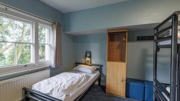 Una cama individual con cabecera, un armario de madera, un radiador, una ventana con cortinas y una cama litera.