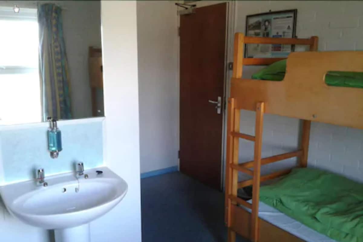 Una cama litera con sábana verde, un lavamanos blanco y un espejo en una habitación con puerta y un cuadro en la pared.