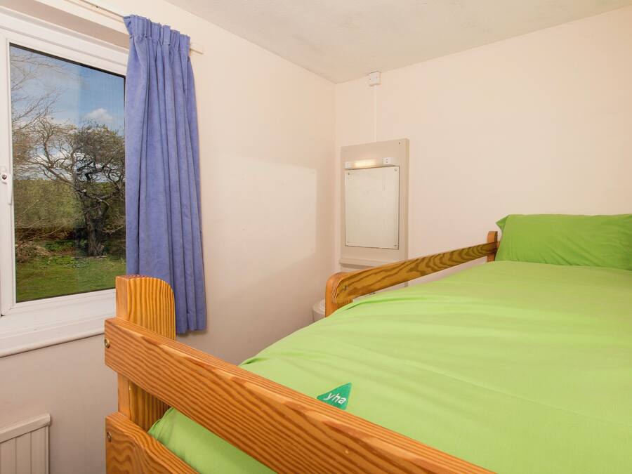 A single bed with a wooden frame and a green mattress in a room with a window and a wall-mounted mirror.