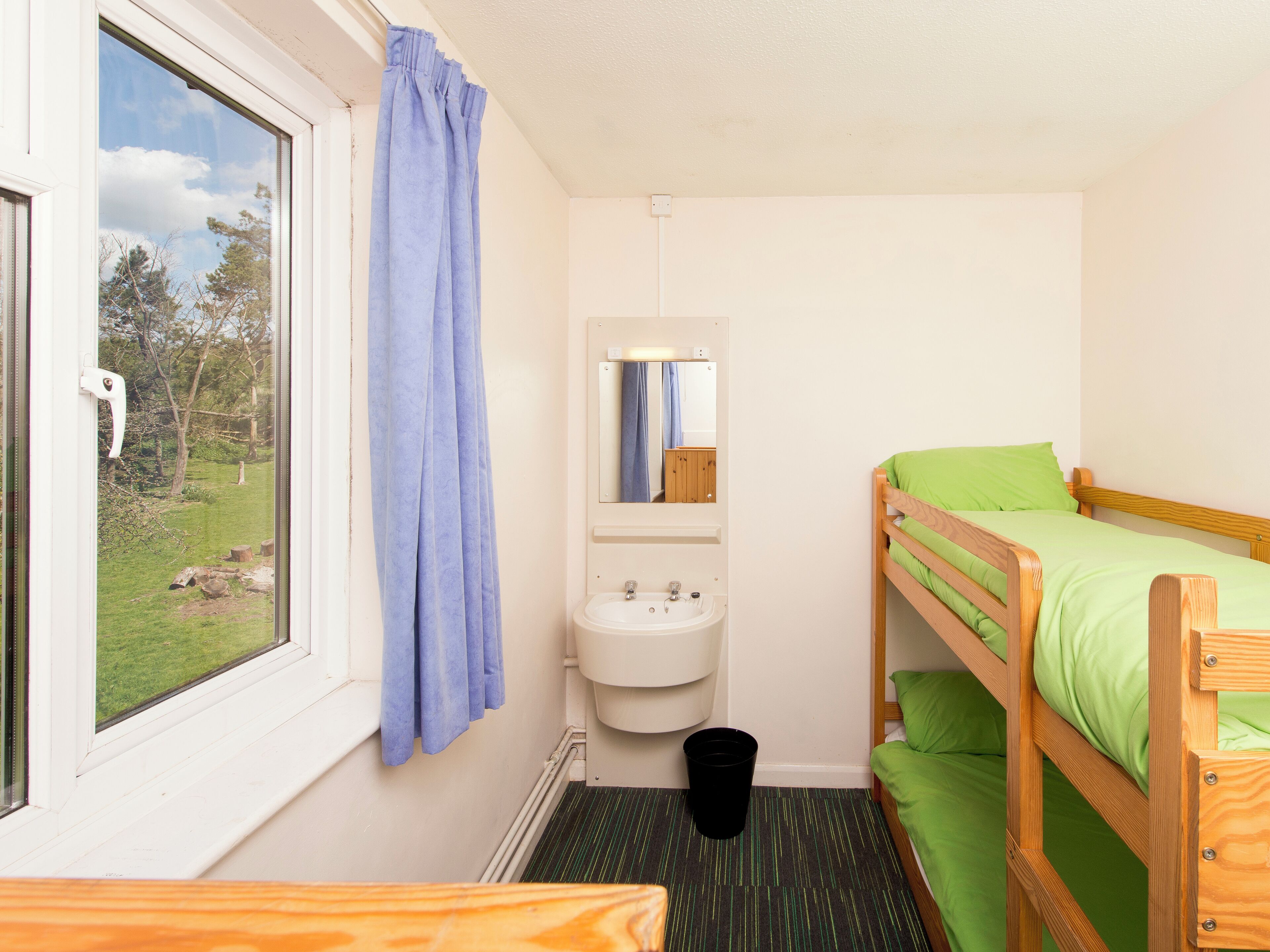 Een kleine, schone kamer met een stapelbed, een raam met uitzicht op bomen, een wastafel en een spiegel.