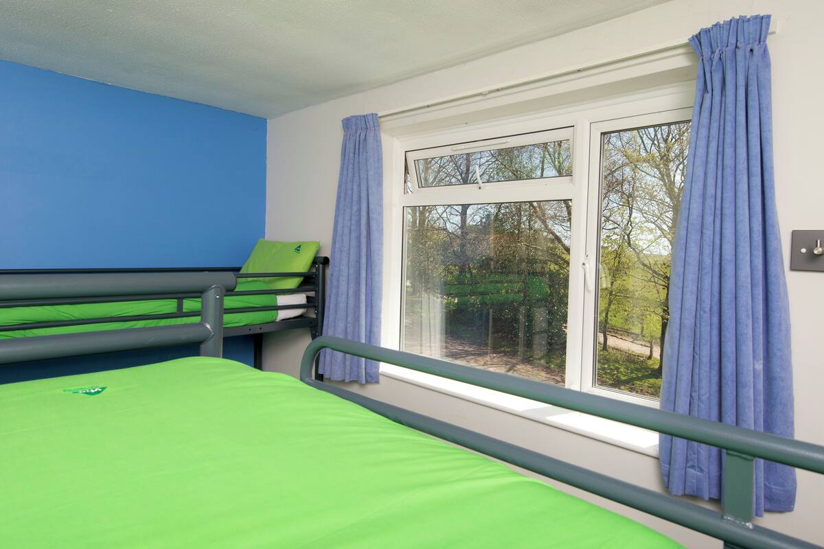 Une chambre avec un lit superposé, une fenêtre avec des rideaux et une vue sur les arbres à l’extérieur.