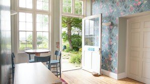 A room with a wooden table, two chairs, a plaid bench, a door with a floral wallpaper, and large windows with a view of greenery.