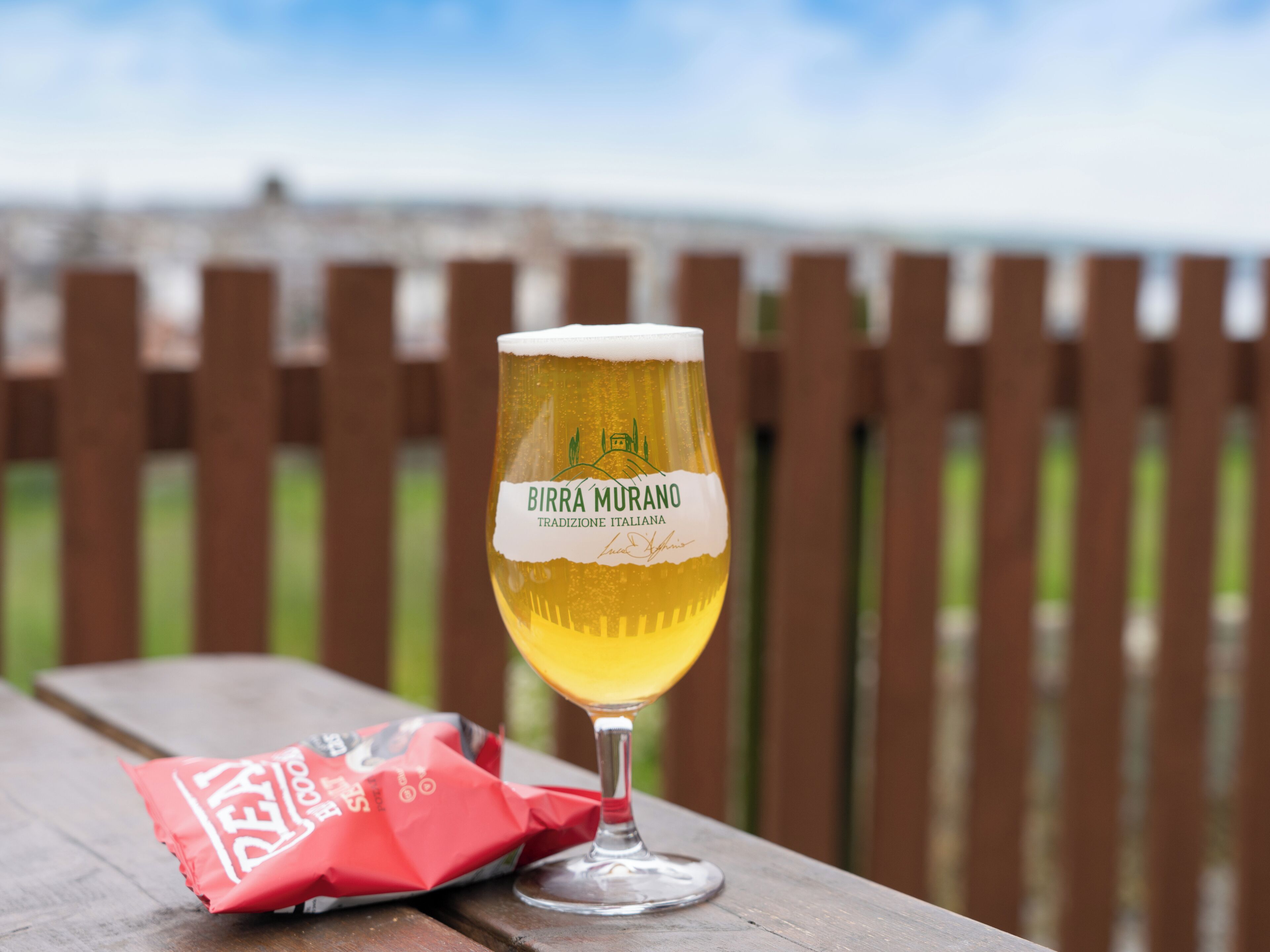 A glass of beer on a wooden table with a blurred background of a fence and sky.