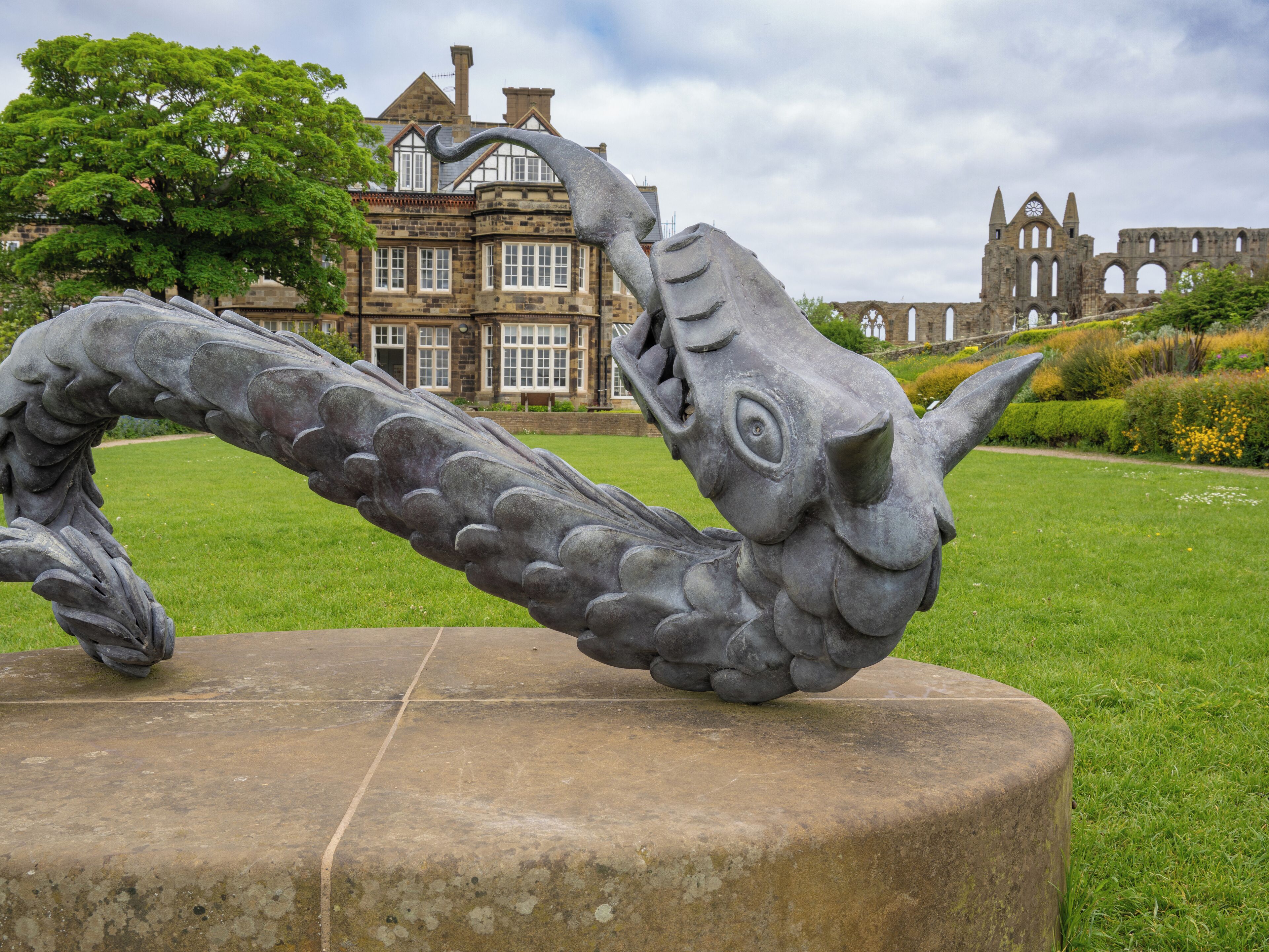 Une sculpture de dragon, posée sur un piédestal en pierre, dans un parc, avec un bâtiment historique en arrière-plan.