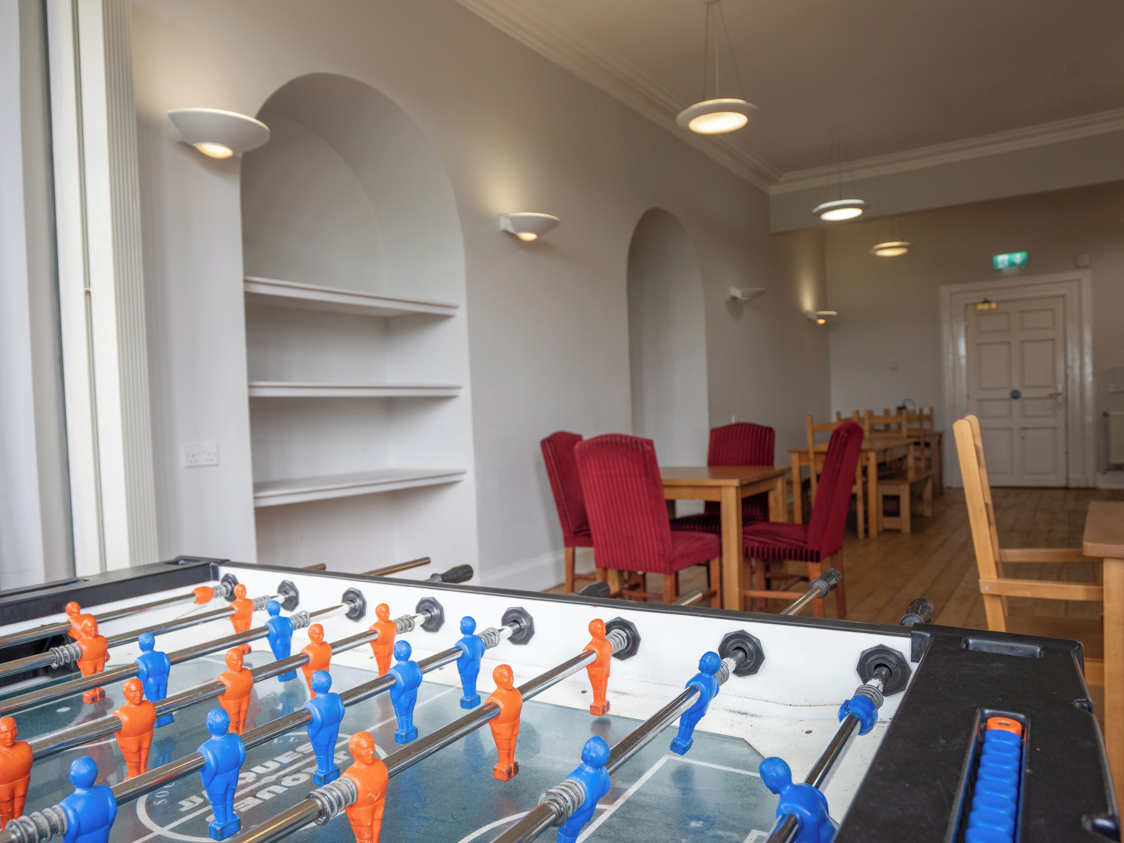 A foosball table in a room with white walls, red chairs, and a wooden table.