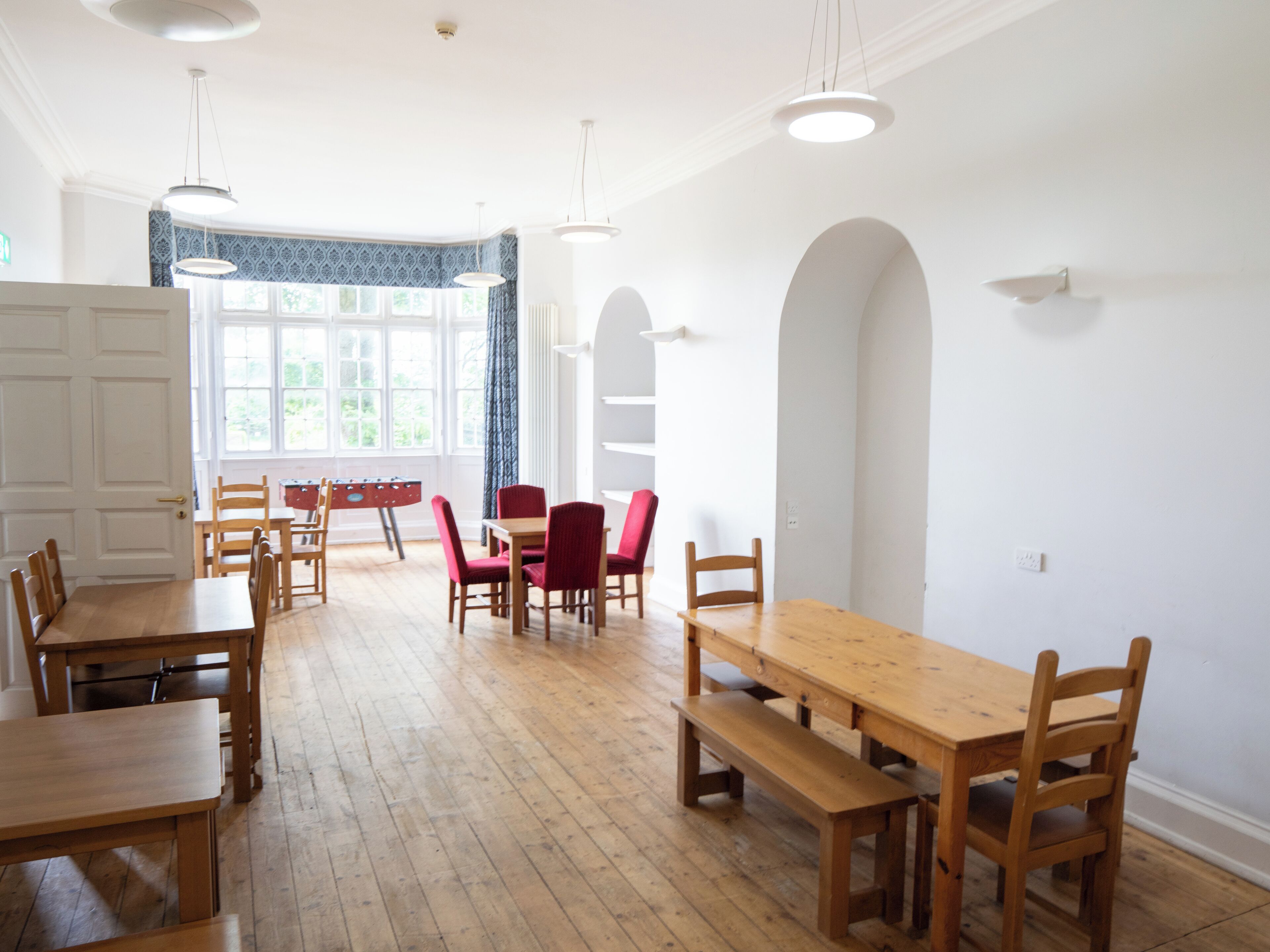 A spacious dining area with wooden tables and chairs, a large window with curtains, and a view of greenery outside.