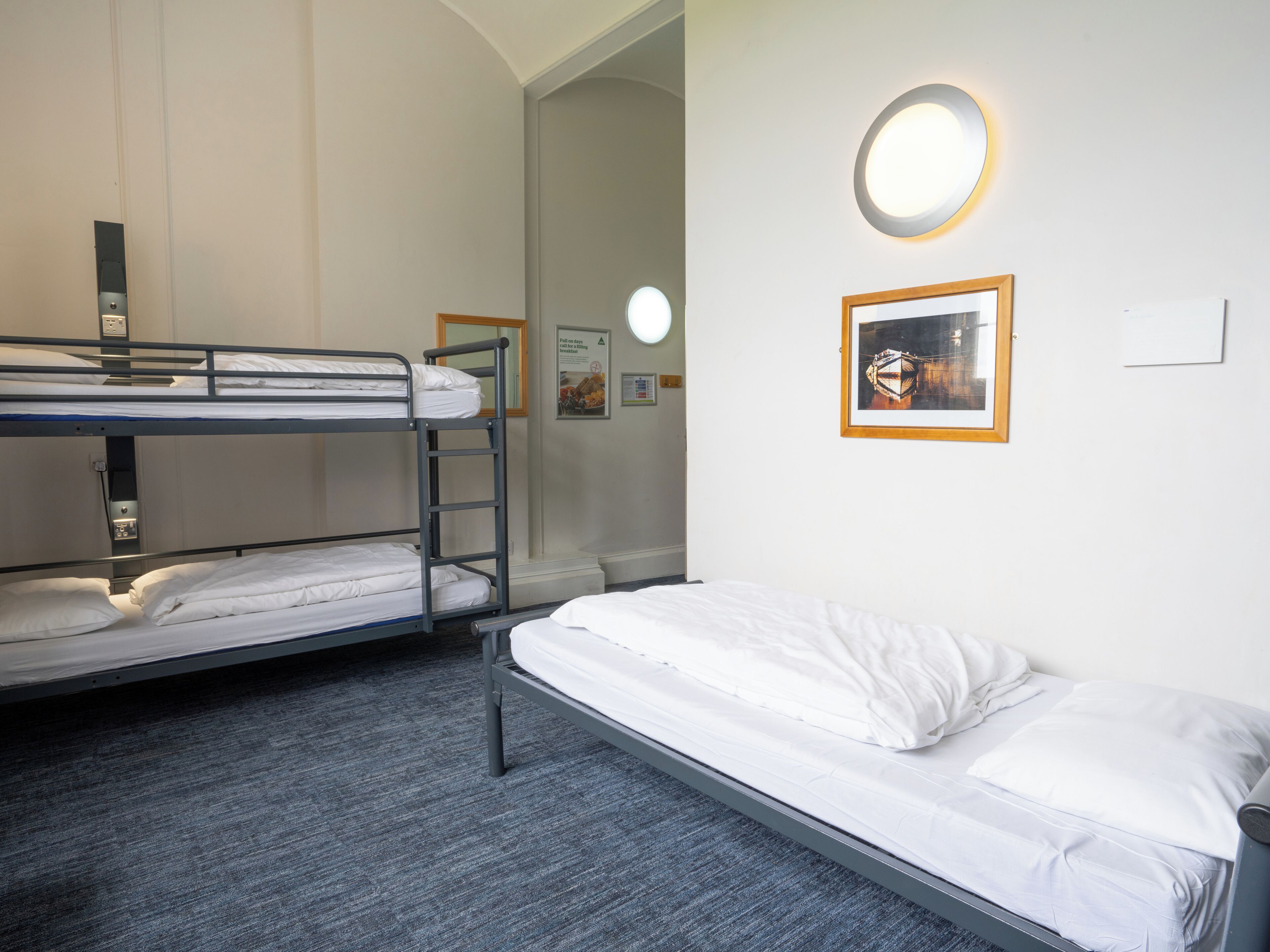 Une chambre d’hôtel avec des lits superposés, un lit simple, un applique murale ronde et un tableau encadré accroché au mur.
