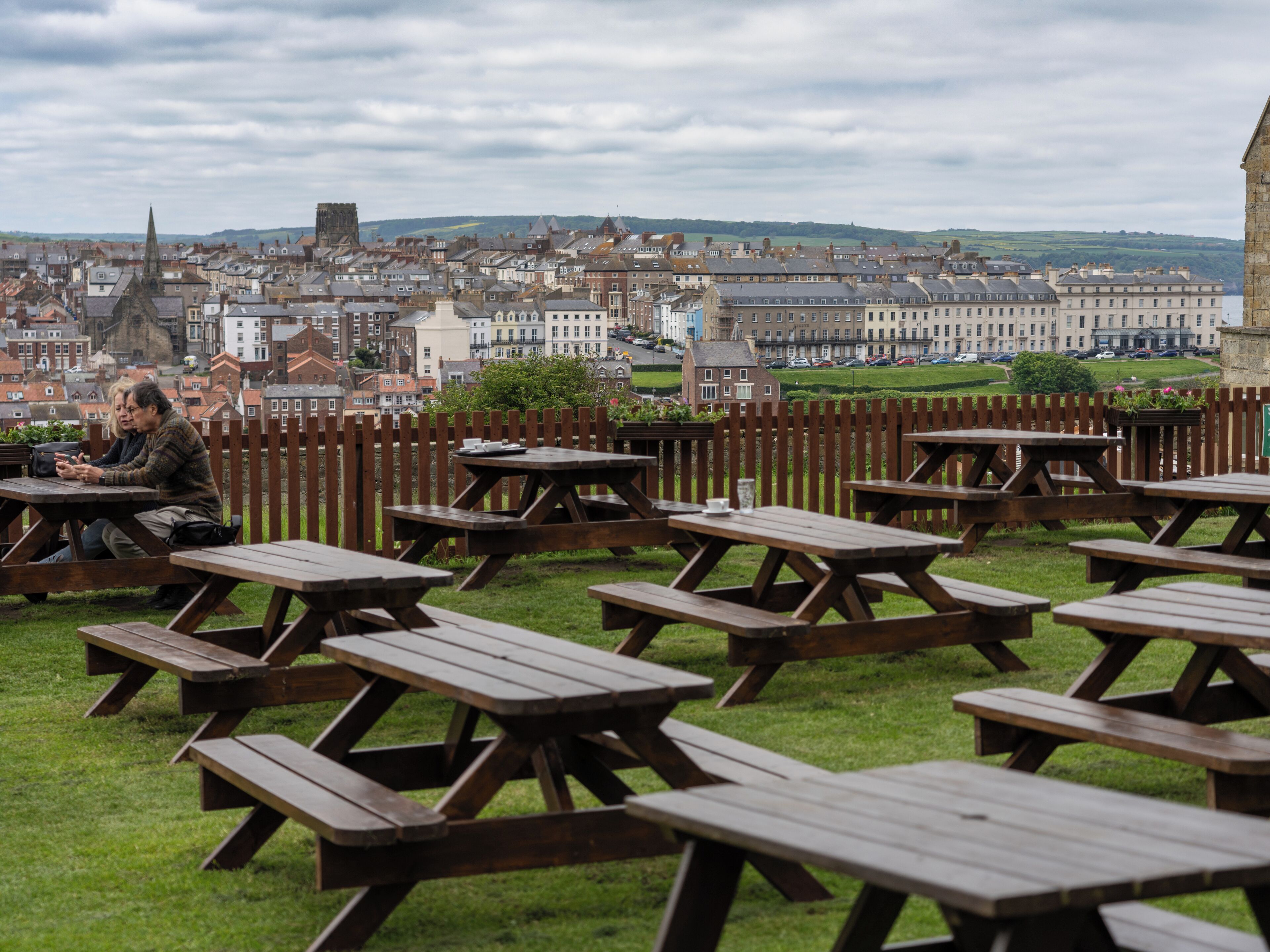 A picnic area with wooden tables and benches, set against a backdrop of a historic town with buildings and a church spire.
