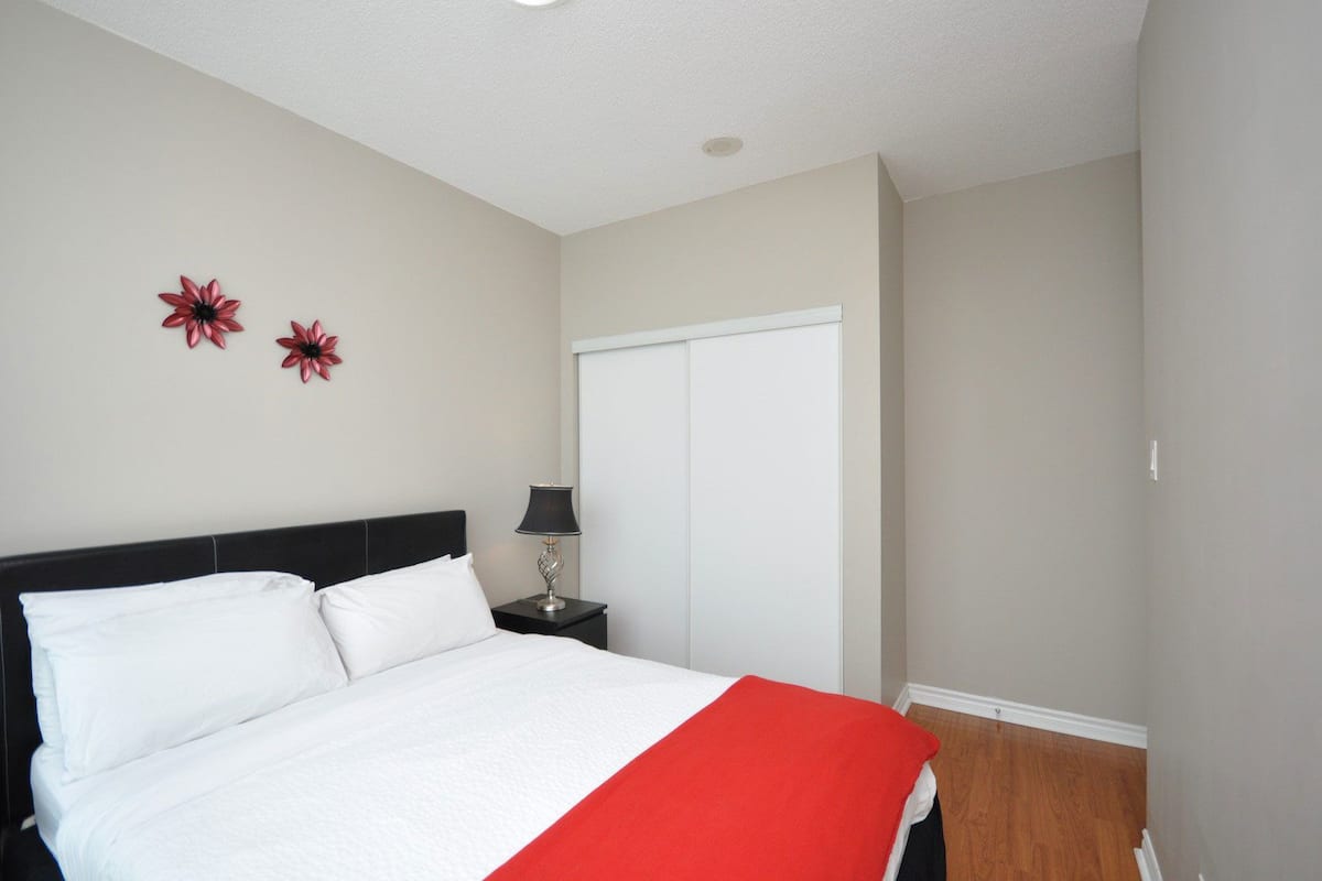 Un lit bien fait, avec une literie blanche et rouge, une table de chevet noire avec une lampe, et une armoire blanche dans une pièce simple aux murs de couleur beige.