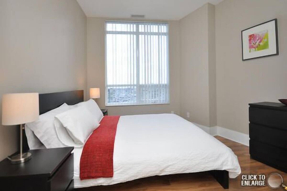 Une chambre à coucher avec un lit, une table de chevet, une lampe, une fenêtre avec des stores et un tableau encadré au mur.